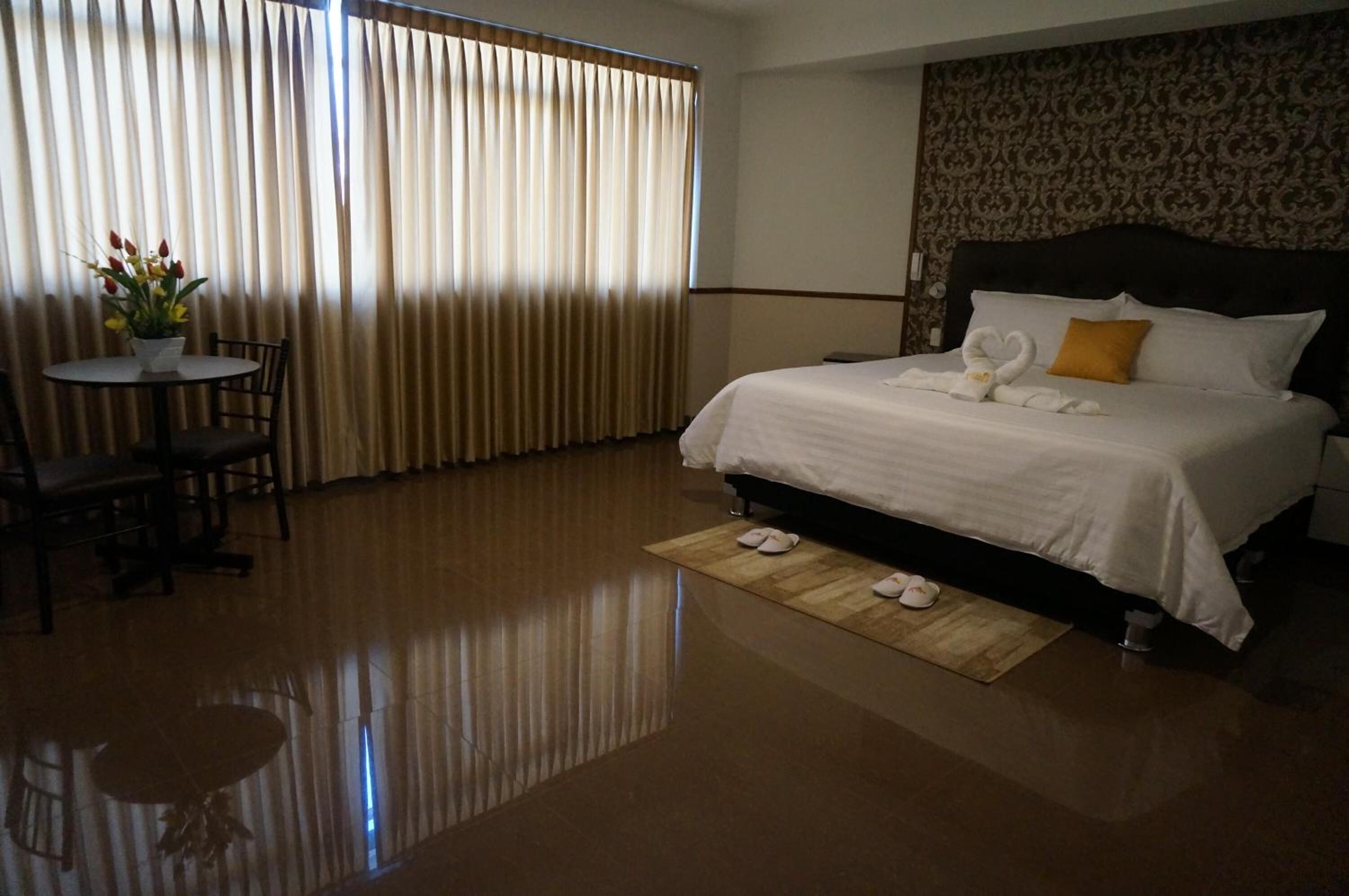 Habitación de hotel con cama, una mesa redonda con sillas, un florero y una ventana con cortinas.