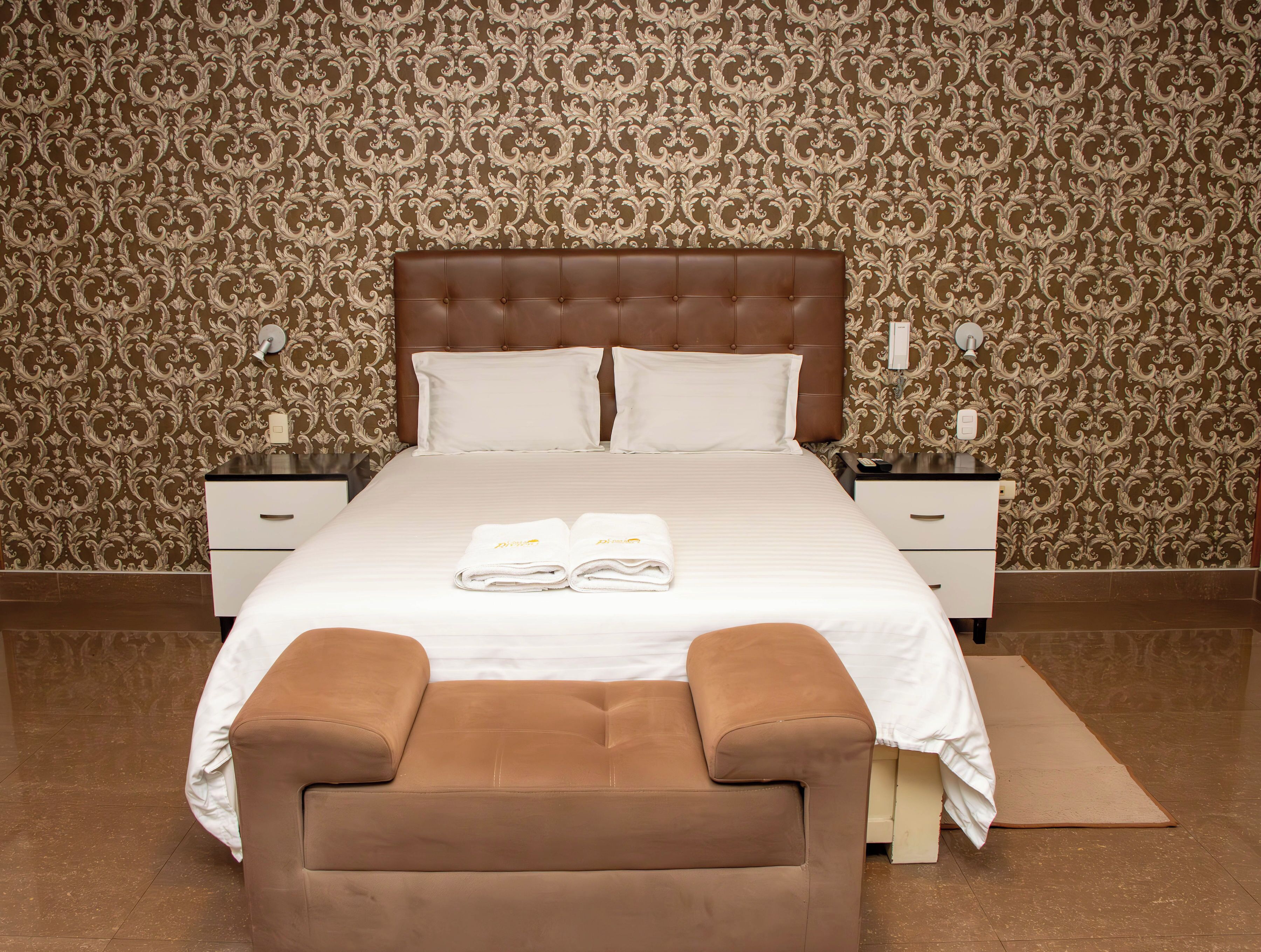 A neatly made bed with white linens, two pillows, and a folded towel on the bed. There are two nightstands with lamps and a telephone on each. A brown upholstered chair is in front of the bed. The room has patterned wallpaper and a brown floor.