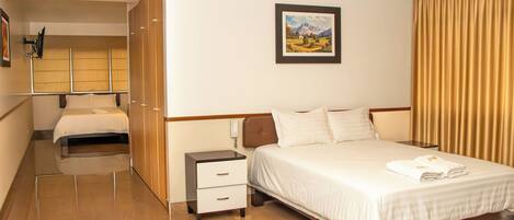 Quarto de hotel com cama, mesas de cabeceira, pequena escrivaninha, cadeira, guarda-roupa e um quadro na parede.