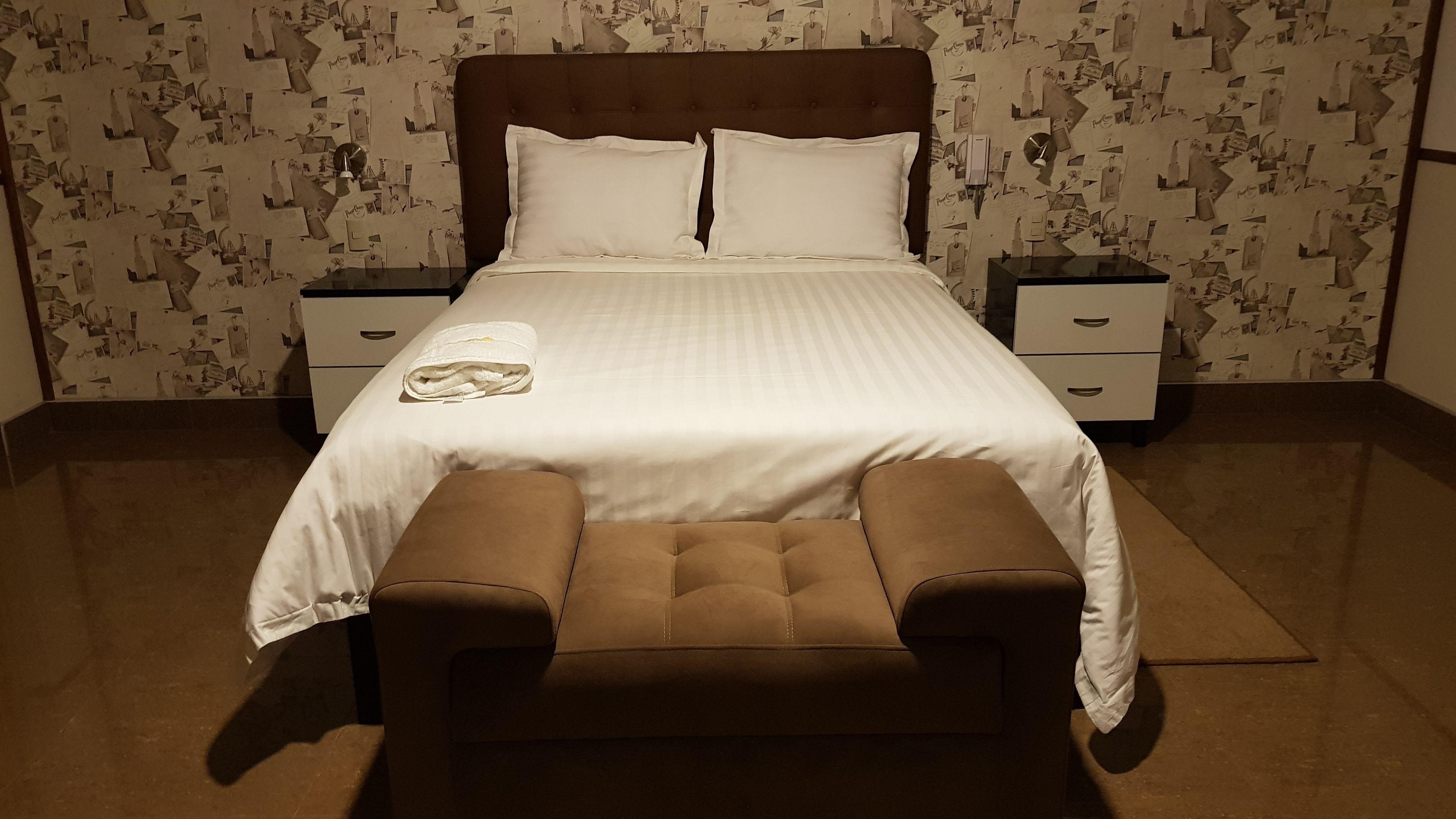 Un llit ben arrenjat amb un estovat blanc i dos coixins, flanquejat per dues taules de nit amb làmpades. L'habitació té un paper pintat i un banc marró al davant del llit.