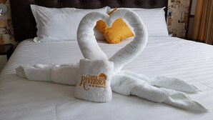 A bed with white linens, a heart-shaped towel arrangement, and a 'Costa Riviera Hotel' towel.