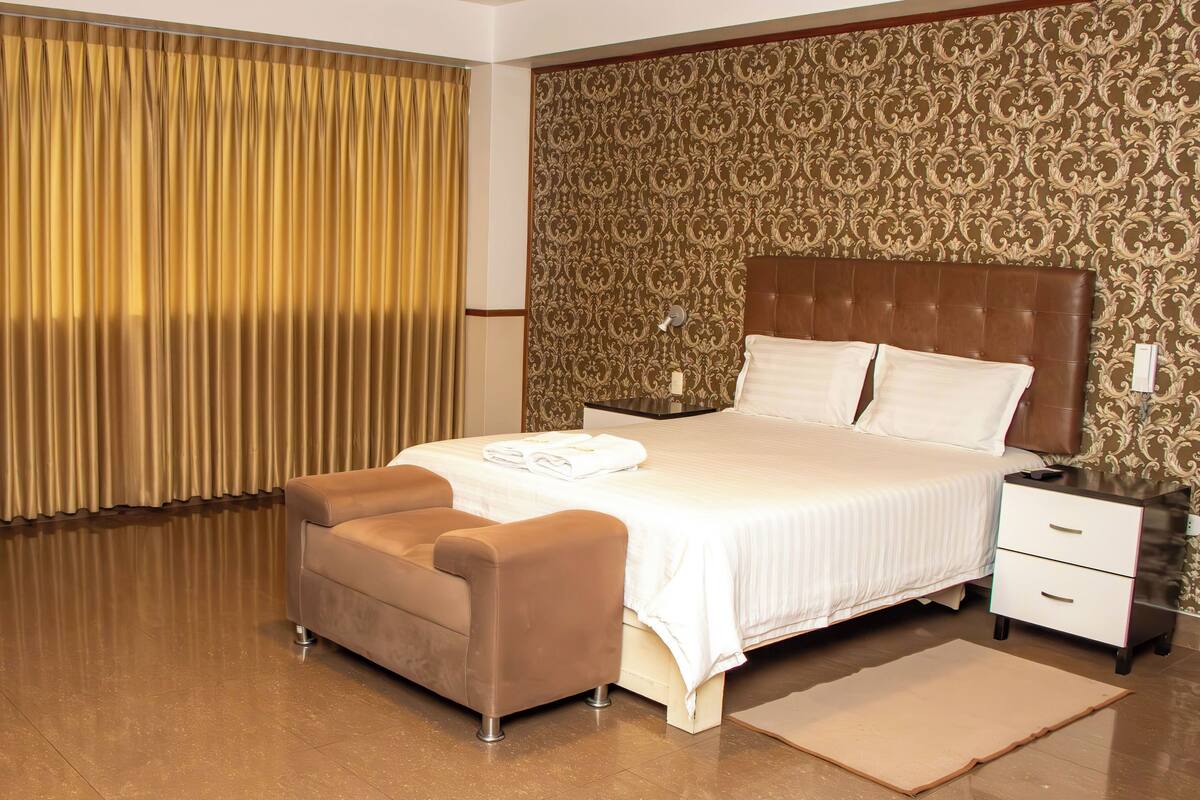 A hotel room with a large bed, a bedside table, a nightstand, a small sofa, a desk with a chair, and a curtain.