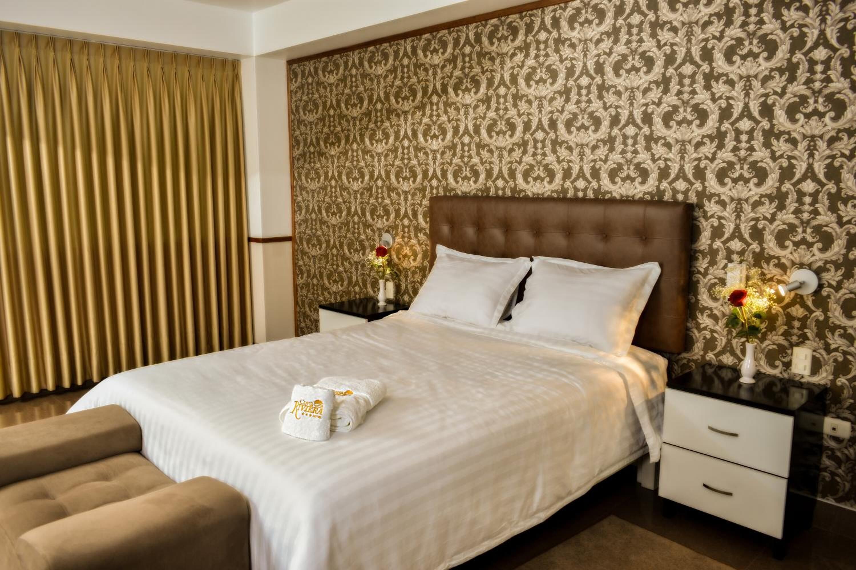 Habitación de hotel con una cama grande, cabecera con estampado floral, mesita de noche con un florero y una ventana con cortinas.