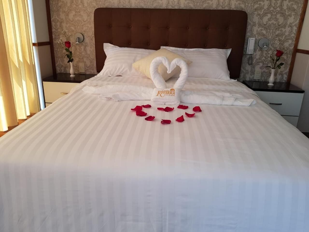 Una cama bien hecha con una toalla con forma de corazón y pétalos de rosa sobre el edredón.