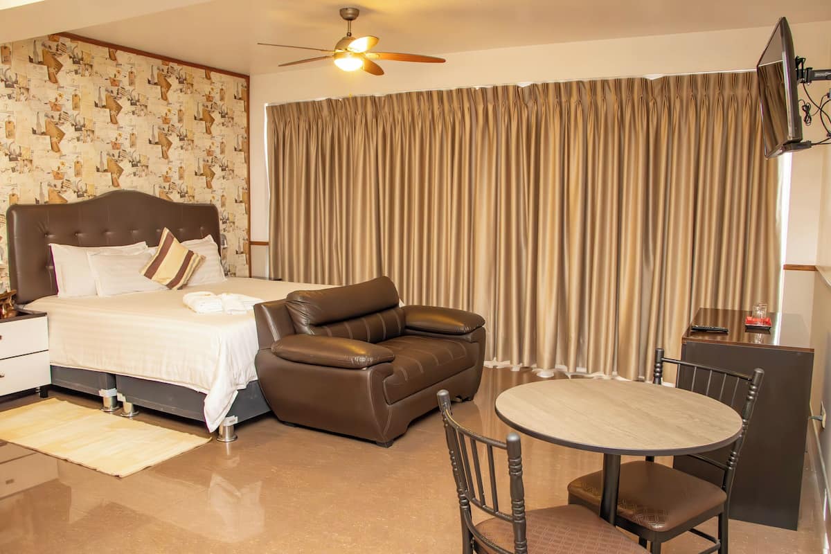 A hotel room with a bed, a leather sofa, a dining table with chairs, a ceiling fan, and a TV mounted on the wall.