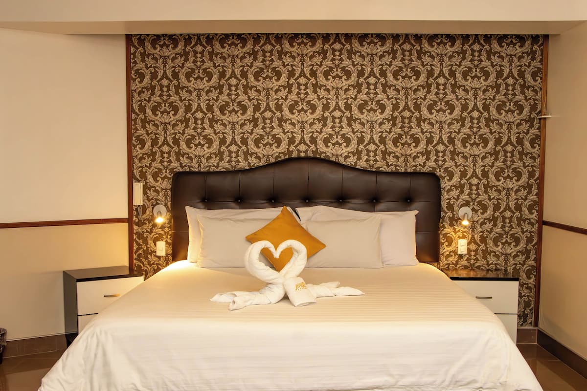 A neatly made bed with a heart-shaped towel and a single pillow, a dark upholstered headboard, and a patterned wallpaper.