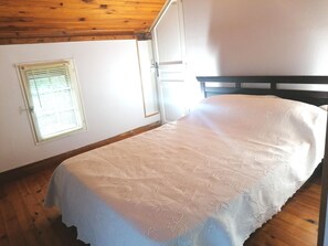 A single bed with a floral-patterned bedspread in a room with wooden ceiling beams and a window with blinds.