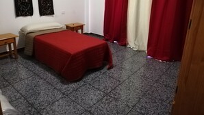 Un dormitorio pequeño y ordenado con una cama, dos mesitas de noche y un ventanal con cortinas rojas.