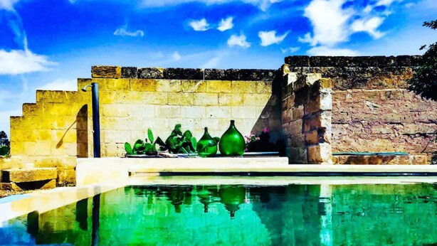 Una piscina bajo un cielo azul claro, con un muro de piedra y jarrones verdes.