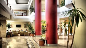 A spacious lobby with high ceilings, red columns, and multiple levels. There are several potted plants and a seating area with people. - Hotel Ephesia (Kuşadası)