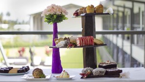 A dessert display with various pastries and cakes on a table, a purple vase with flowers, and a balcony railing in the background. - Crossroads Hotel Lae (Lae)