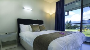 A neatly made bed with a white comforter and a brown throw blanket, a bedside table with a lamp, and a large window with a view of a green landscape. - Crossroads Hotel Lae (Lae)