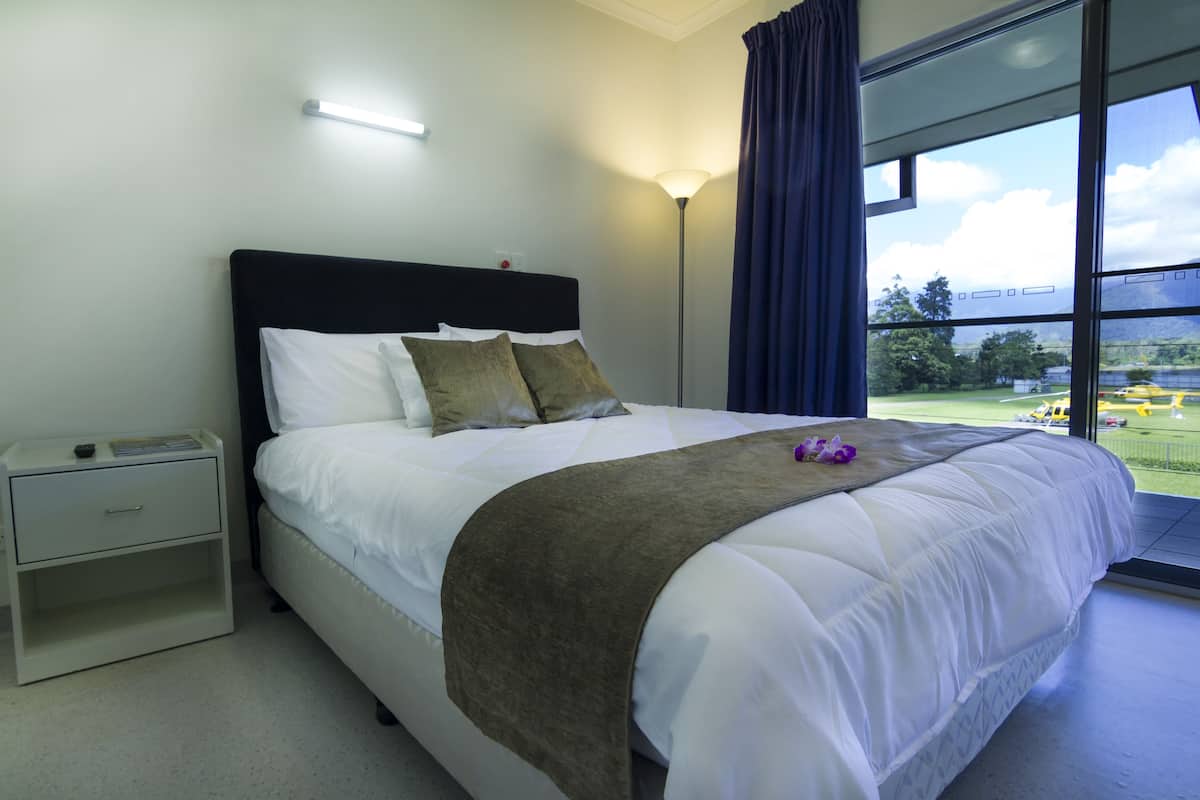 A neatly made bed with a white comforter and a brown throw blanket, a bedside table with a lamp, and a large window with a view of a green landscape.