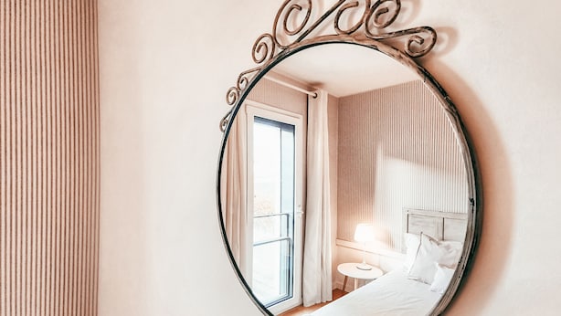 Een hotelkamer met een grote spiegel in ovaalvorm, een bed met witte lakens, een nachtkastje met een lamp, en een weergave van het interieur van de kamer in de spiegel.