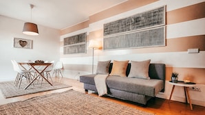 Een moderne woonkamer met een bank, een eettafel met stoelen en een groot raam met zonwering.