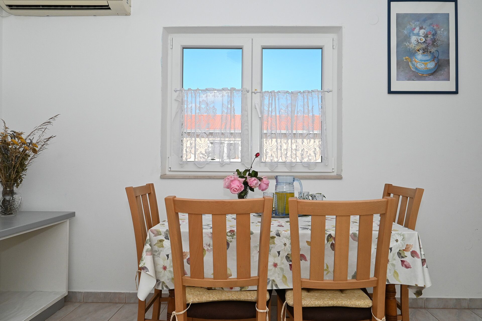 A dining area with a table set for four, a window with lace curtains, and a vase with flowers on the left.