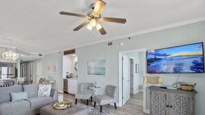 Un salon moderne avec un canapé gris, des fauteuils à motifs, un ventilateur de plafond et un téléviseur posé sur un meuble.