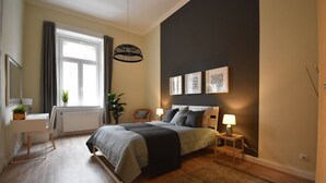 Un dormitorio con cama, mesitas de noche, escritorio, silla, radiador, ventana con cortinas y decoración en la pared.