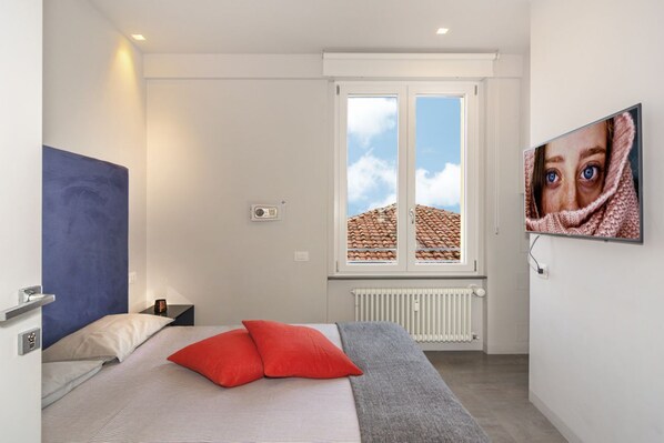 A bedroom with a bed, a window with a view of a tiled roof, a radiator, and a wall-mounted TV. - Mediceo 42 - Standard Double Room (Pisa)