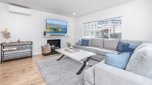 A modern living room with a grey sofa, a flat-screen TV, a fireplace, and a large window overlooking a balcony.