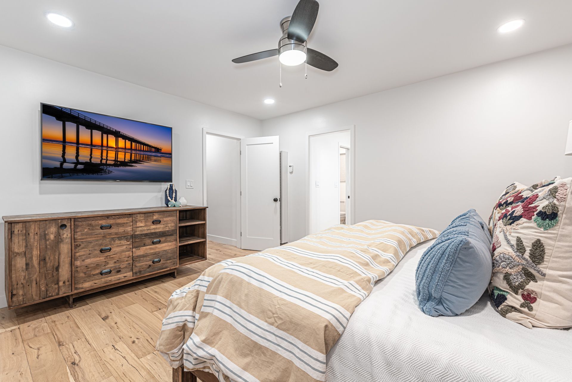 Een slaapkamer met een houten dressoir, een bed met gestreept bedlinnen, een plafondventilator en een schilderij van een pier.