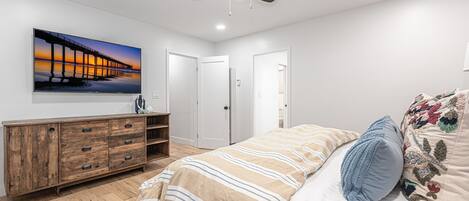 Een slaapkamer met een houten dressoir, een bed met gestreept bedlinnen, een plafondventilator en een schilderij van een pier.