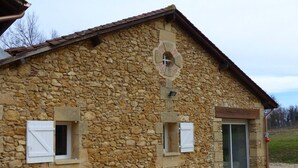 A stone building with a sloped roof, two small windows with shutters, and a door with a glass panel. - GITE VERY COMFORTABLE IN THE COUNTRY, NICE VOLUMES PROMOTION IN JULY (Cours-de-Pile)