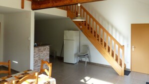 A spacious room with a wooden staircase, a dining area with a table and chairs, a kitchen with a refrigerator, and a door leading to another room. - GITE VERY COMFORTABLE IN THE COUNTRY, NICE VOLUMES PROMOTION IN JULY (Cours-de-Pile)