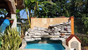 A swimming pool with a stone waterfall, surrounded by tropical plants and a wooden structure. - Casa Leyani y Osvedy (Viñales)