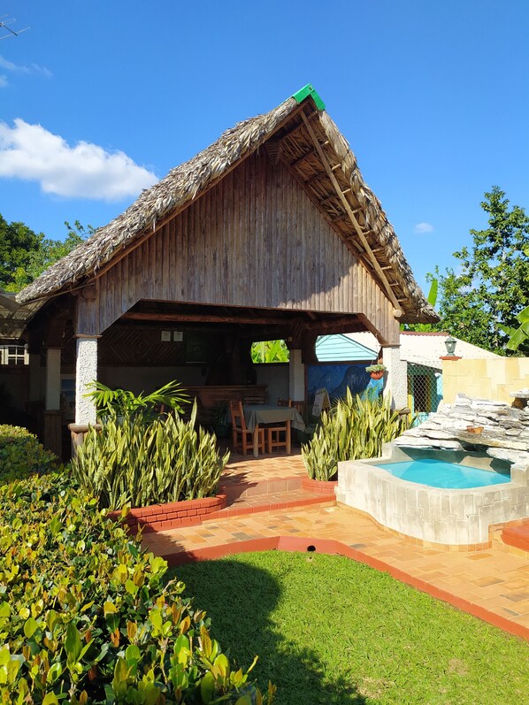 A rustic wooden building with a thatched roof, a dining area with a table and chairs, and a small pool with a stone waterfall.