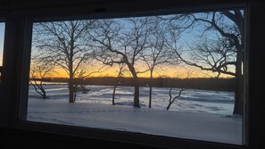 Vista a través de una ventana de un paisaje nevado con árboles desnudos y un atardecer.