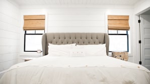Une chambre avec un grand lit, deux fenêtres équipées de stores en bois, un plafond blanc et une table de chevet en bois.