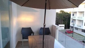 Un balcon avec une table et des chaises, un parasol, et une vue sur la rue et les immeubles.