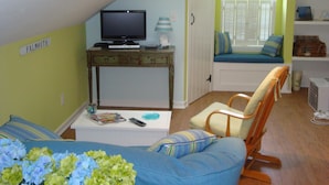A cozy living room with a television on a wooden table, a comfortable chair, and a window seat with a view of greenery. - Quaint Studio near Mashpee Commons (East Falmouth)