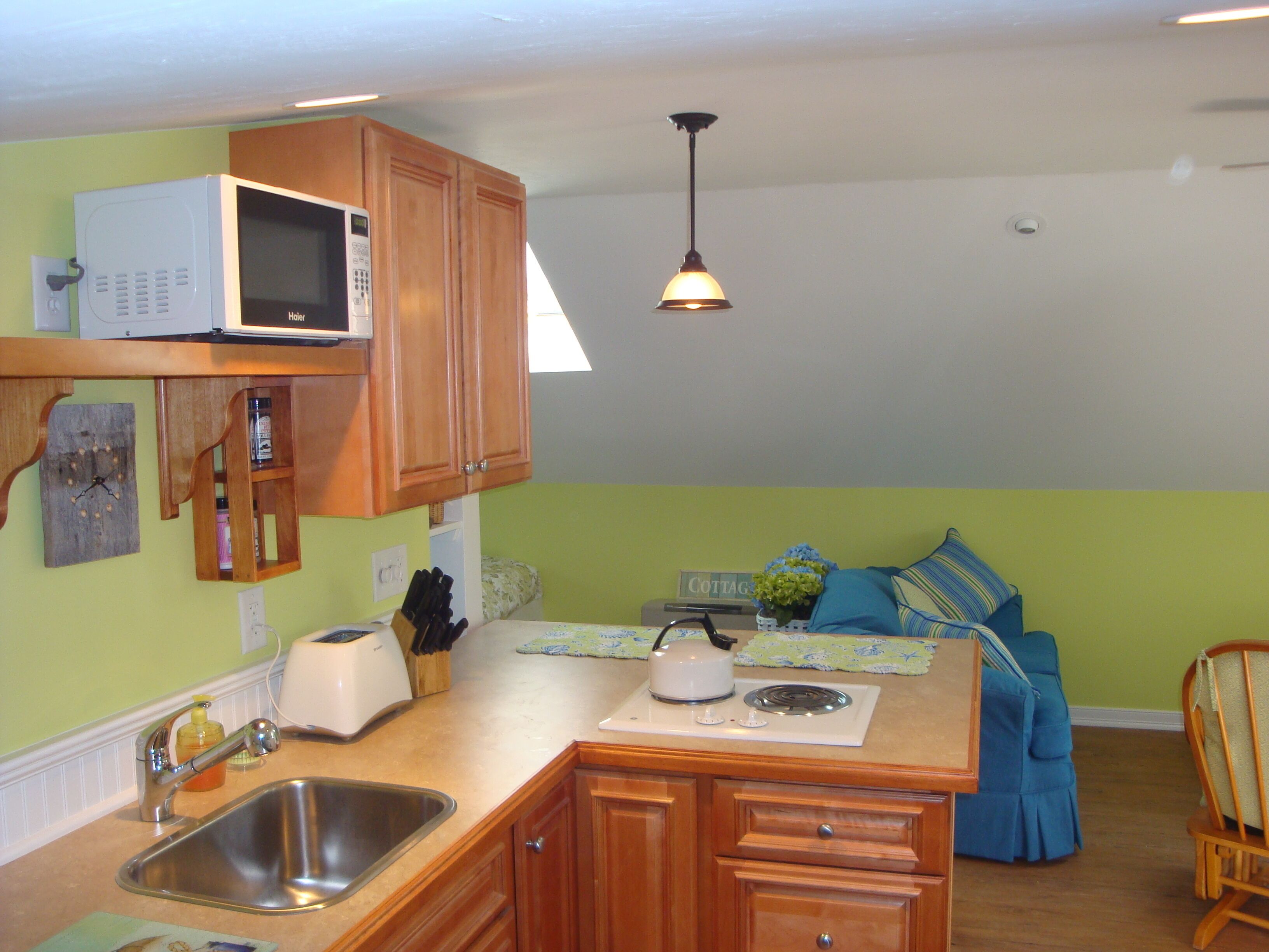 A compact kitchen with a microwave, sink, and wooden cabinets. There is a small island with a kettle and a toaster. The room has a green wall and a ceiling light.