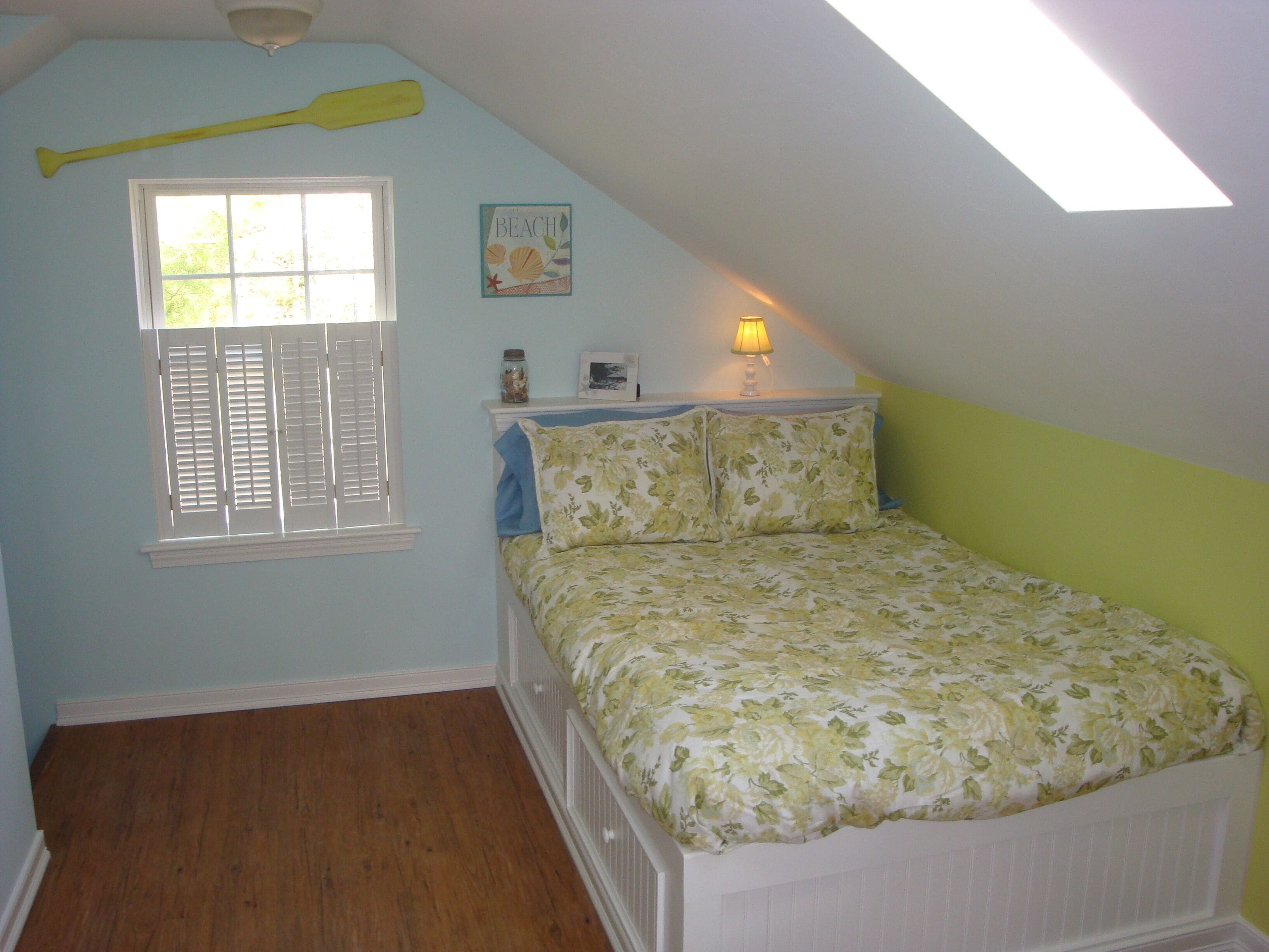 Ein Schlafzimmer mit schrägverdachter Decke, einem Fenster mit Fensterläden, einem Bett mit blumigem Bettbezug, einem Nachttisch mit Lampe und einer Wanddekoration.