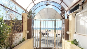 A gated entrance to a beach area with a covered walkway leading to the beach, a bench, and a dog on the path.