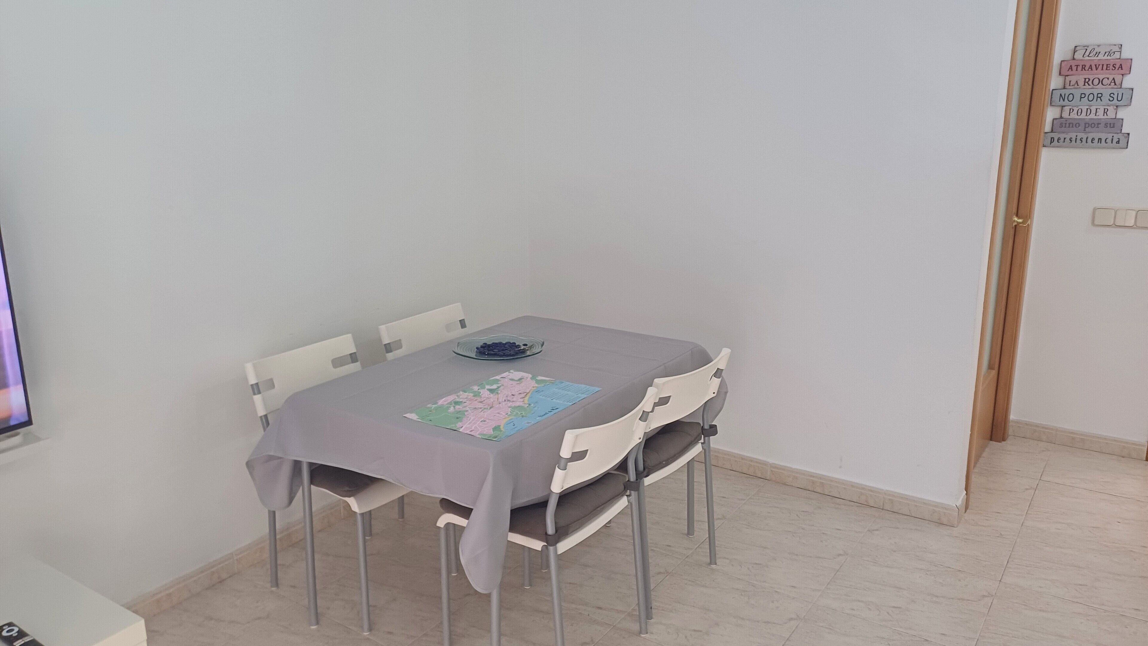 A dining area with a table covered by a tablecloth, chairs, a bowl of fruit, and a wall with a motivational quote.