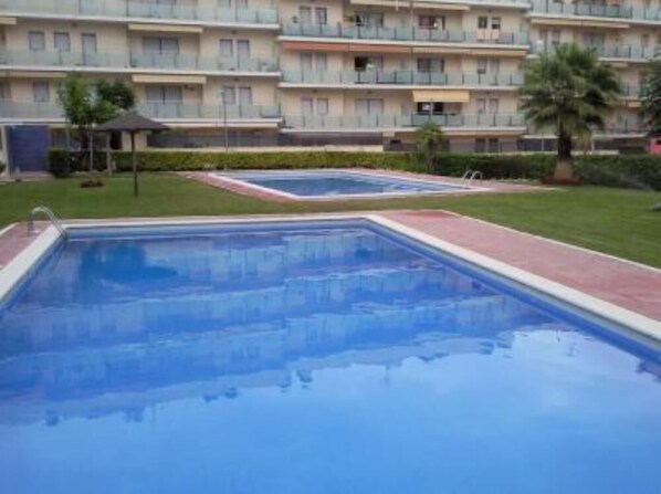 A swimming pool with a tiled deck, surrounded by a well-maintained lawn and a multi-story building with balconies.