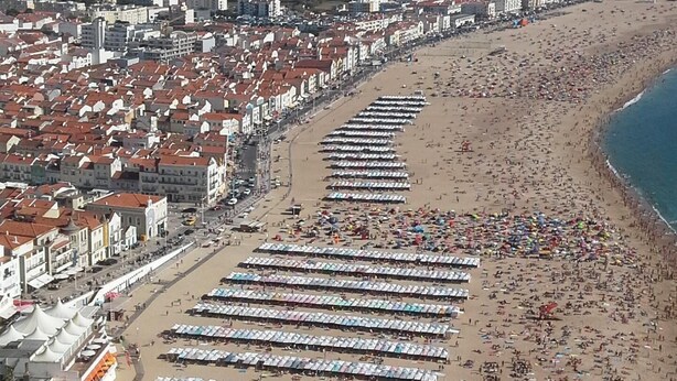 A coastal cityscape with a long beach lined with rows of beach umbrellas and tents. - Villa for rent in Nazaré Portugal (Nazaré)