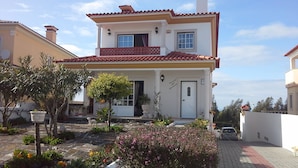 A two-story house with a red-tiled roof, a balcony with a lattice railing, and a front door with a small porch area. - Villa for rent in Nazaré Portugal (Nazaré)