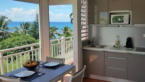 Un balcón con vistas al océano y palmeras, una mesa de comedor con platos y un frutero, y una kitchenette con microondas y fregadero.