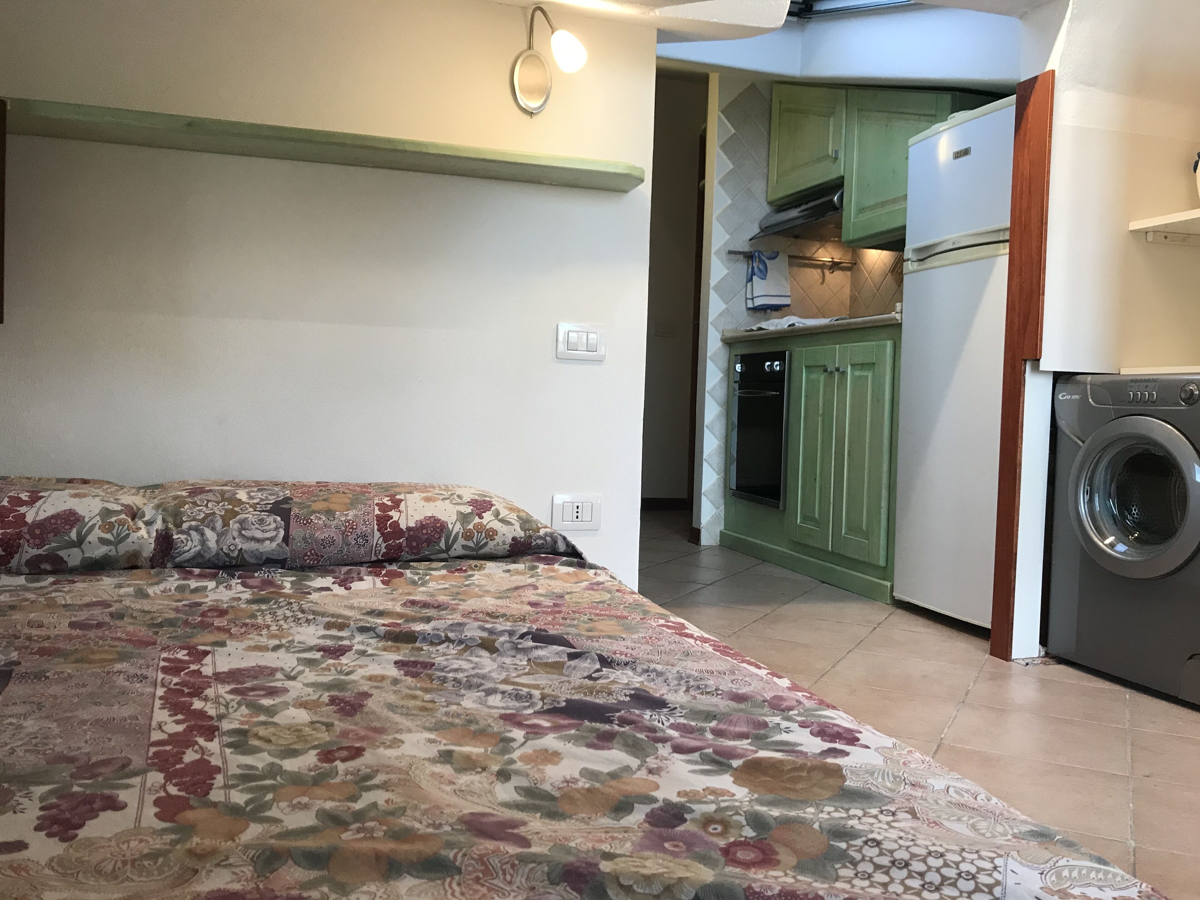 Un dormitorio con colcha estampada, una cocina verde con lavadora y un pequeño comedor.