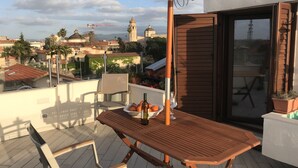 Une table en bois avec une bouteille et des verres, une chaise et une plante en pot sur un balcon d’où l’on aperçoit des bâtiments et un dôme au loin.