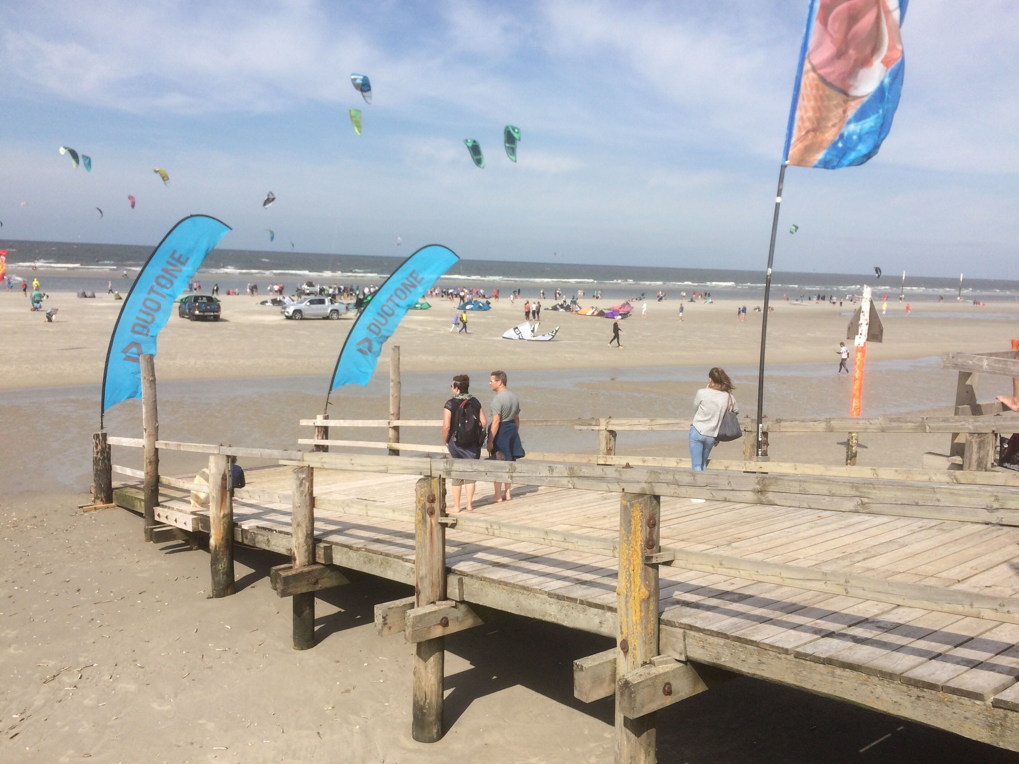 A beach with a wooden pier, flags, and people walking.
