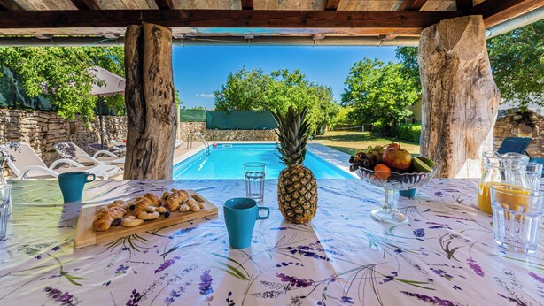 Un área de comedor al aire libre con vista a la piscina, vigas de madera y una mesa dispuesta con comida y bebidas.
