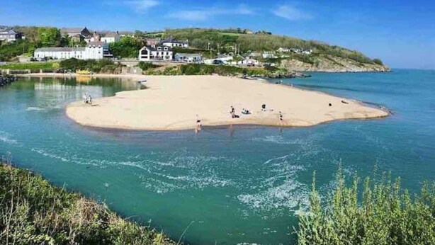 Uma praia costeira com águas claras, areal arenoso e casas ao fundo.