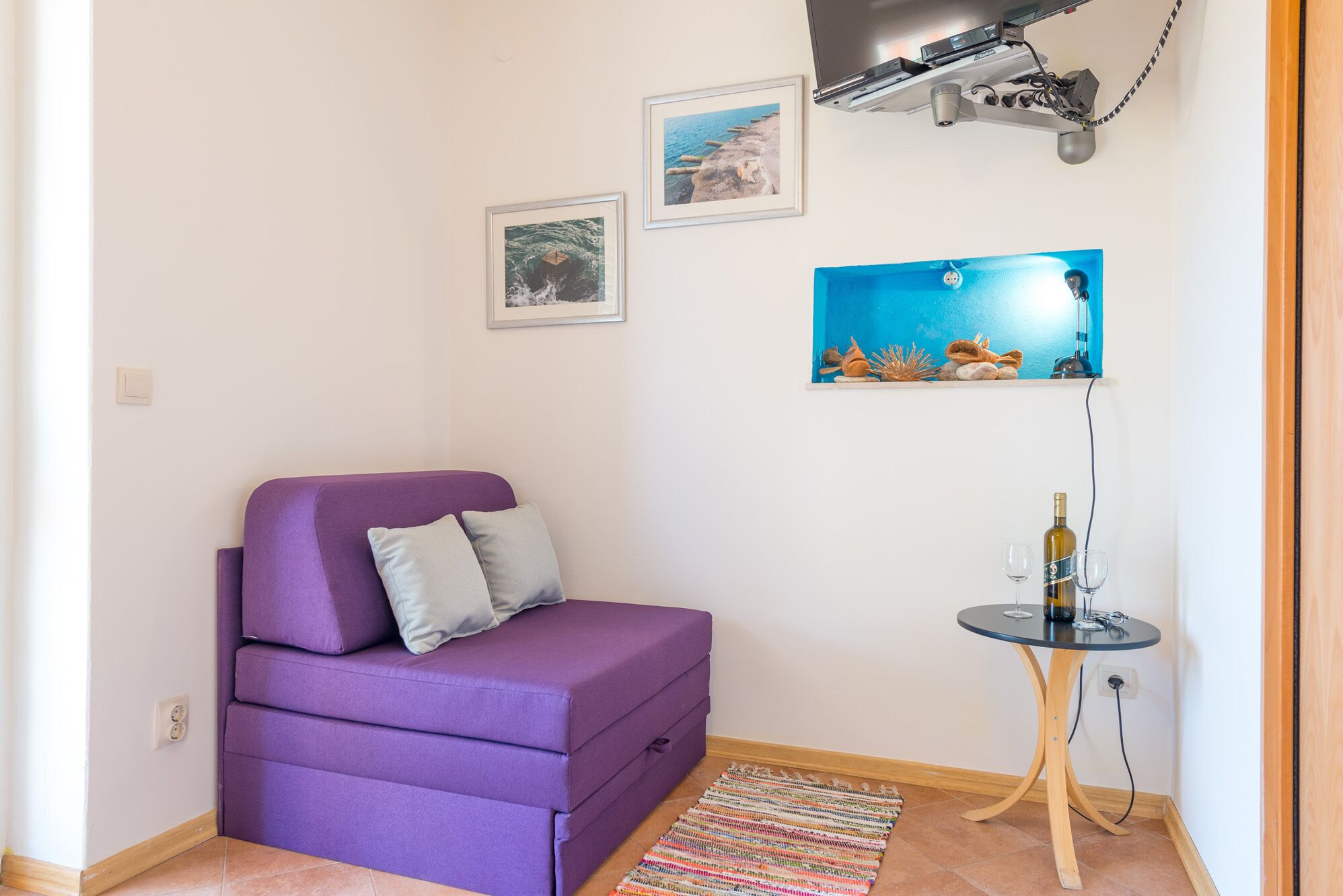 A small room with a purple sofa, a small table with a bottle and glasses, and two framed pictures on the wall.