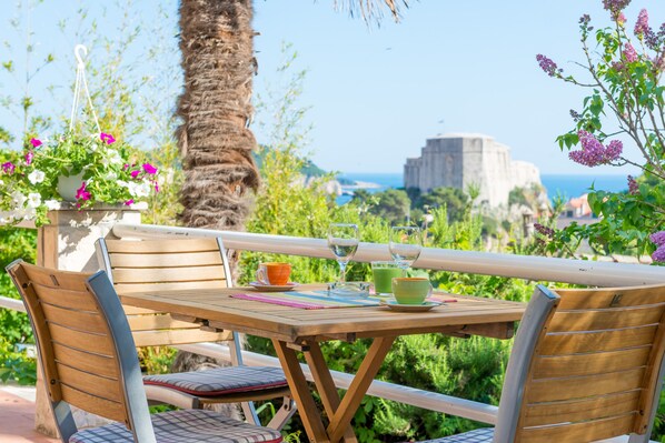 An outdoor seating area with wooden furniture, a table with a placemat, and a view of the sea and a historic building in the background.