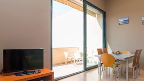 Un salon moderne avec un téléviseur à écran plat posé sur un meuble en bois, un coin repas avec une table et des chaises, et une grande porte-fenêtre coulissante donnant sur un balcon extérieur.